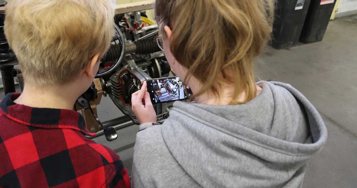 Kaksi opiskelijaa tutkii moottoria ja ottaa kuvaa kännykällä