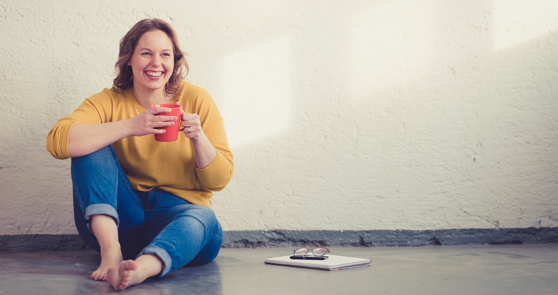 "Hymyilevä nainen istuu lattialla kahvikuppi käsissään"