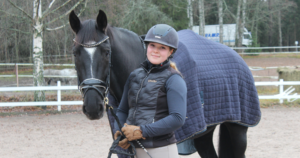 Ratsastusasussa oleva nainen hymyilee ja seisoo mustan hevosen vieressä.