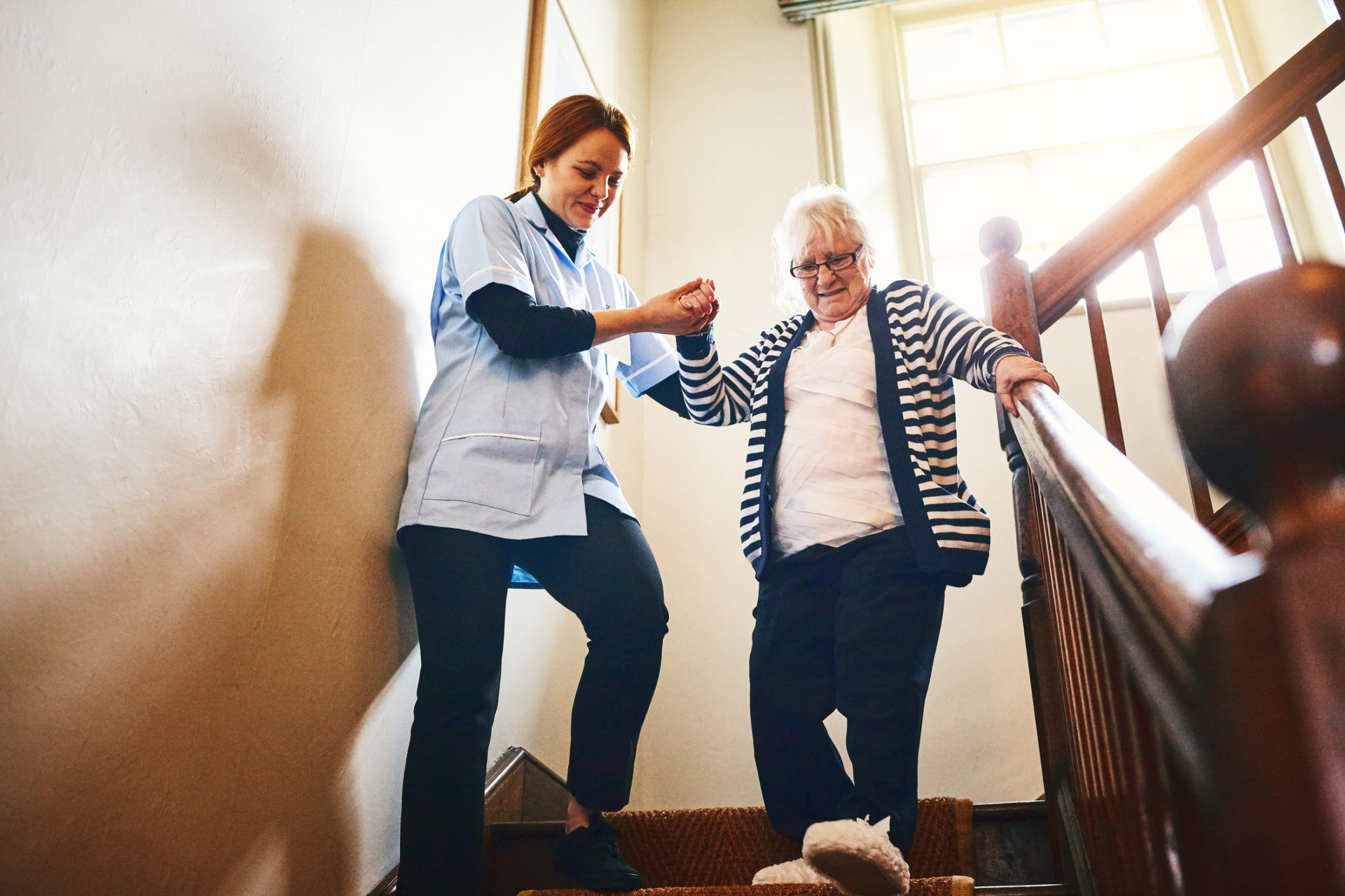 Young female caregiver helping senior woman walking down stairs at home