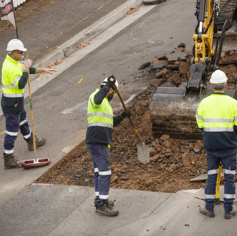 Kolme henkilöä tekee tien korjaustyötä kaivurin avustamana.