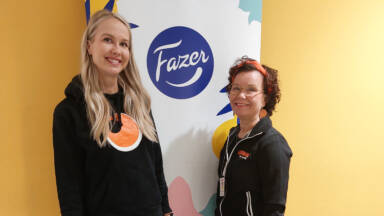 Two women are standing in front of a sign with the Fazer logo. One is blonde and wearing a black hoodie with an orange logo. The other has curly hair, glasses, a red headband, and a black zip-up jacket. The background is yellow.