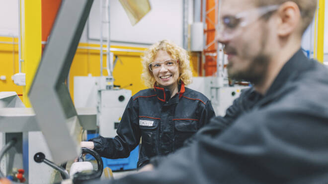 A woman with curly blonde hair and glasses smiles while working on machinery in a factory. She is wearing protective gear. A man in the foreground, also in protective gear, focuses on operating equipment. Bright yellow walls are in the background.