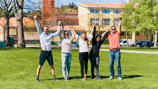 A group of five people standing on a green lawn on a sunny day, holding hands and raising them triumphantly in the air. They are smiling and appear to be celebrating or enjoying the moment. Behind them are trees, parked cars, and buildings with yellow facades and red rooftops, suggesting a school or campus setting.