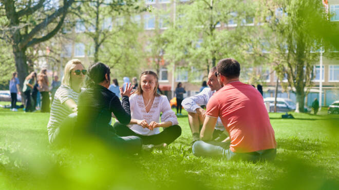 A group of people is sitting in a circle on the grass in a park, engaged in conversation. Trees and buildings are visible in the background on a sunny day. One person is wearing a white shirt and another a red shirt.