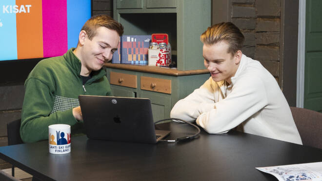 Two people sitting at a table with a laptop and mug, smiling and looking at the screen. A colorful presentation is displayed on a TV in the background. The room has wooden walls and a cabinet with decorative items.