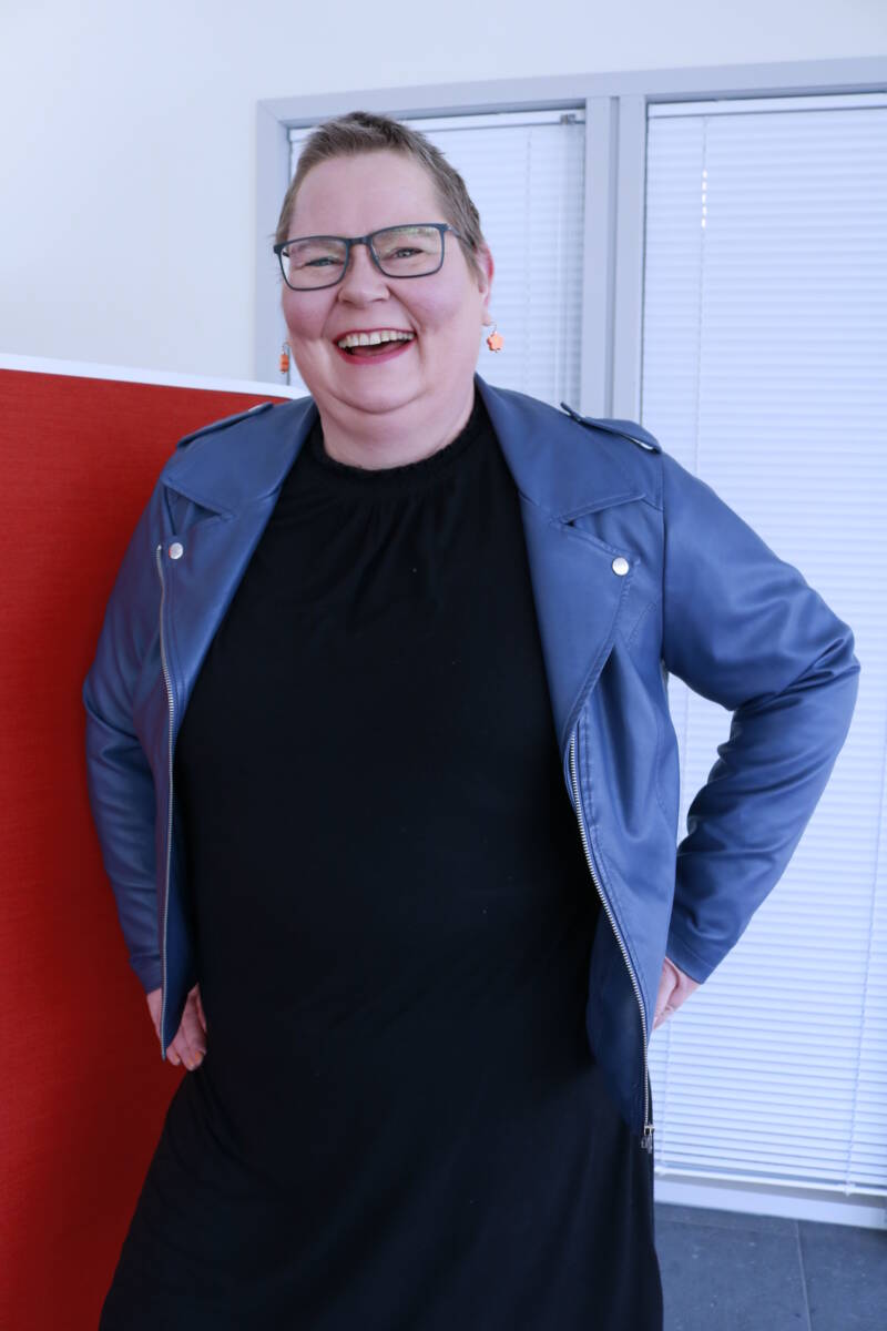 Lyhythiuksinen ja silmälasillinen henkilö seisoo hymyillen kädet lanteilla. Heillä on yllään sininen takki mustan mekon päällä sisätiloissa, joissa on valkoiset kaihtimet ja punainen väliseinä takana.