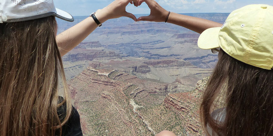 Kaksi tyttöä istuvat korkean vuoren huipulla ja yhdistävät kätensä sydämen muotoiseksi.