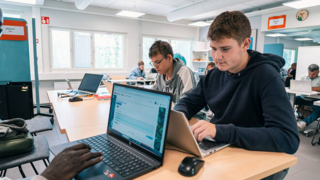 Oppilaat keskittyivät kannettaviin tietokoneisiin istuessaan luokkahuoneessa työpöydän ääressä. Huone on valoisa ja siinä on suuret ikkunat, ja muut opiskelijat näkyvät taustalla. Tunnelma on ahkera ja teknologiavetoinen.