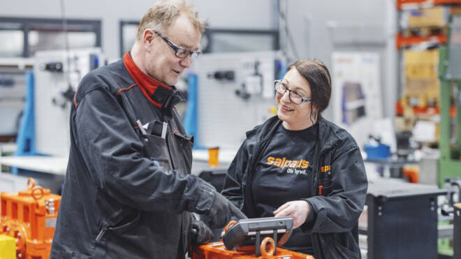 Kaksi henkilöä suojalaseissa ja työtakeissa keskustelevat teollisessa ympäristössä. He seisovat oranssinväristen koneiden lähellä, ja taustalla näkyy hyllyjä ja laitteita.