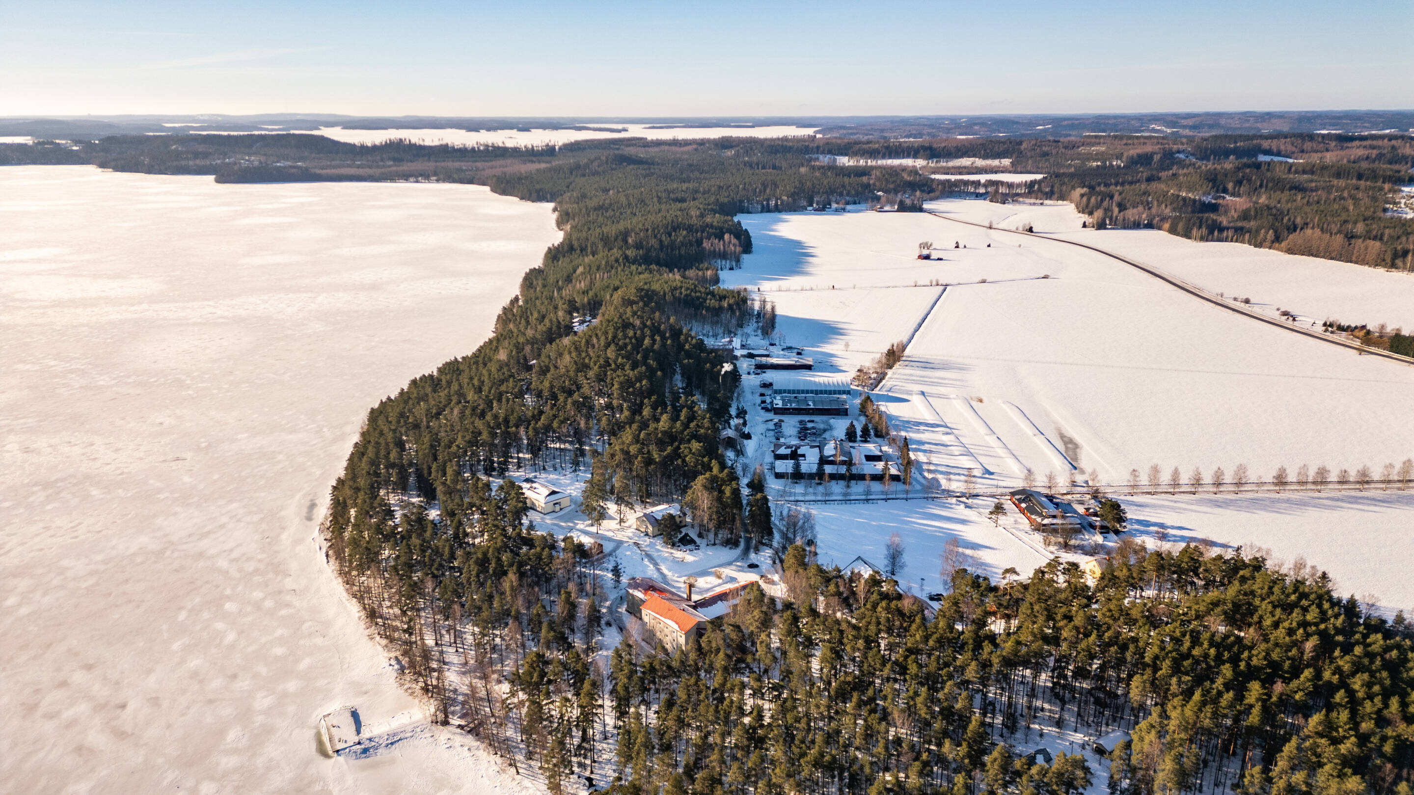 Ilmakuva lumisesta maisemasta, jossa on tiheä metsä, jota ympäröi jäätynyt vesi. Muutamia rakennuksia on hajallaan puiden läheisyydessä, ja taustalla on lumen peittämiä peltoja kirkkaan sinisen taivaan alla.