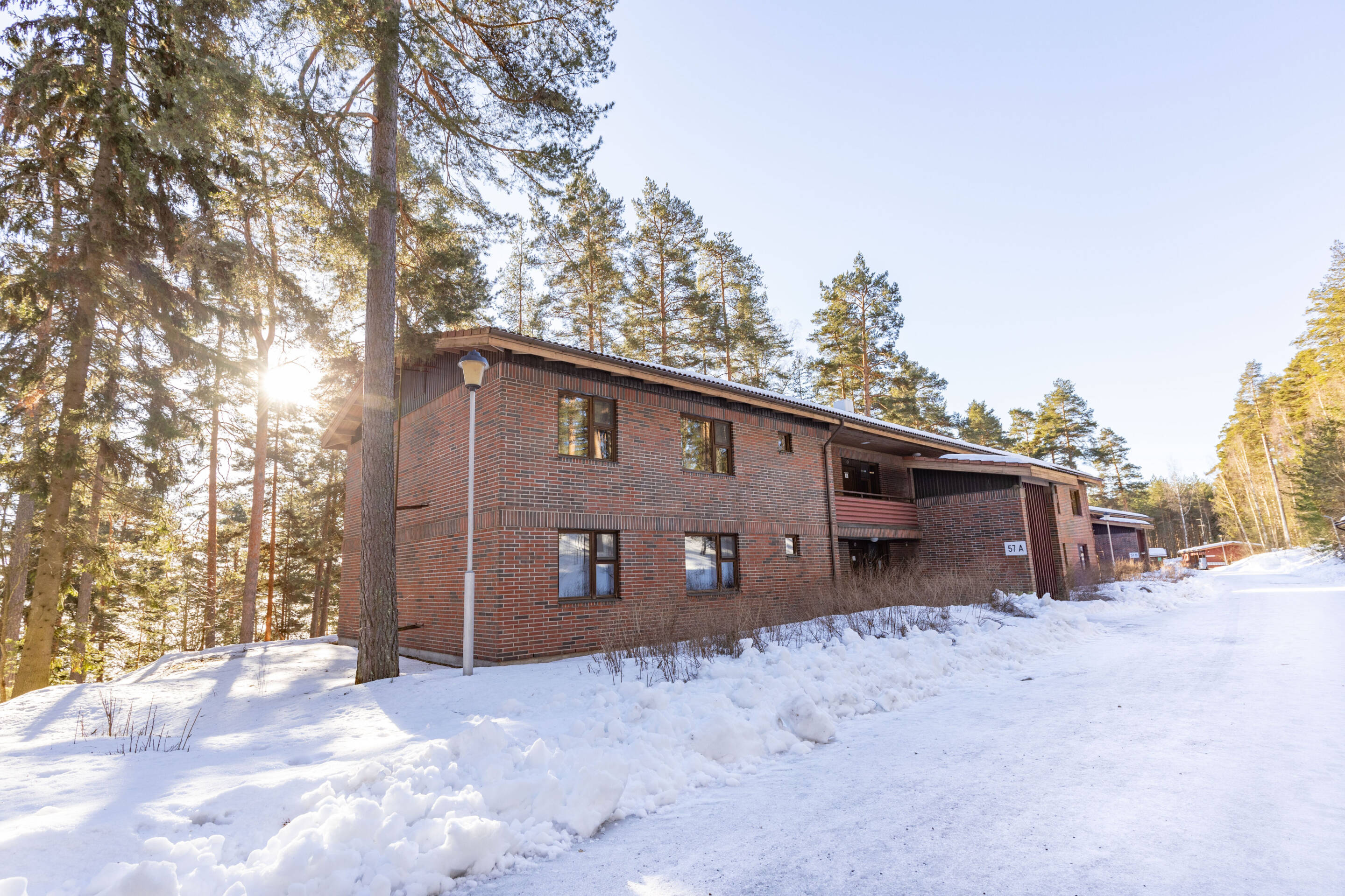 Kaksikerroksinen tiilirakennus lumisessa maisemassa, korkeiden mäntyjen ympäröimänä. Aurinko paistaa puiden läpi luoden varjoja lumen peittämälle maalle. Taivas on kirkas ja sininen.