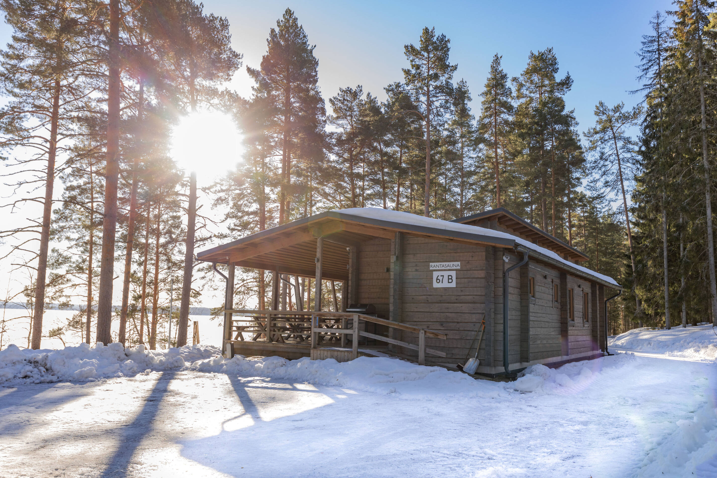 Puinen mökki, jonka nimi on 67 B, seisoo korkeiden mäntyjen ja lumen ympäröimänä. Aurinko paistaa kirkkaasti puiden läpi luoden pitkiä varjoja lumiselle maalle.