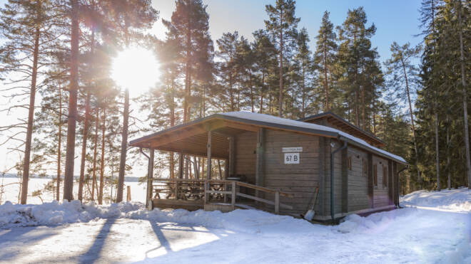 Puinen mökki, jonka nimi on 67 B, seisoo korkeiden mäntyjen ja lumen ympäröimänä. Aurinko paistaa kirkkaasti puiden läpi luoden pitkiä varjoja lumiselle maalle.
