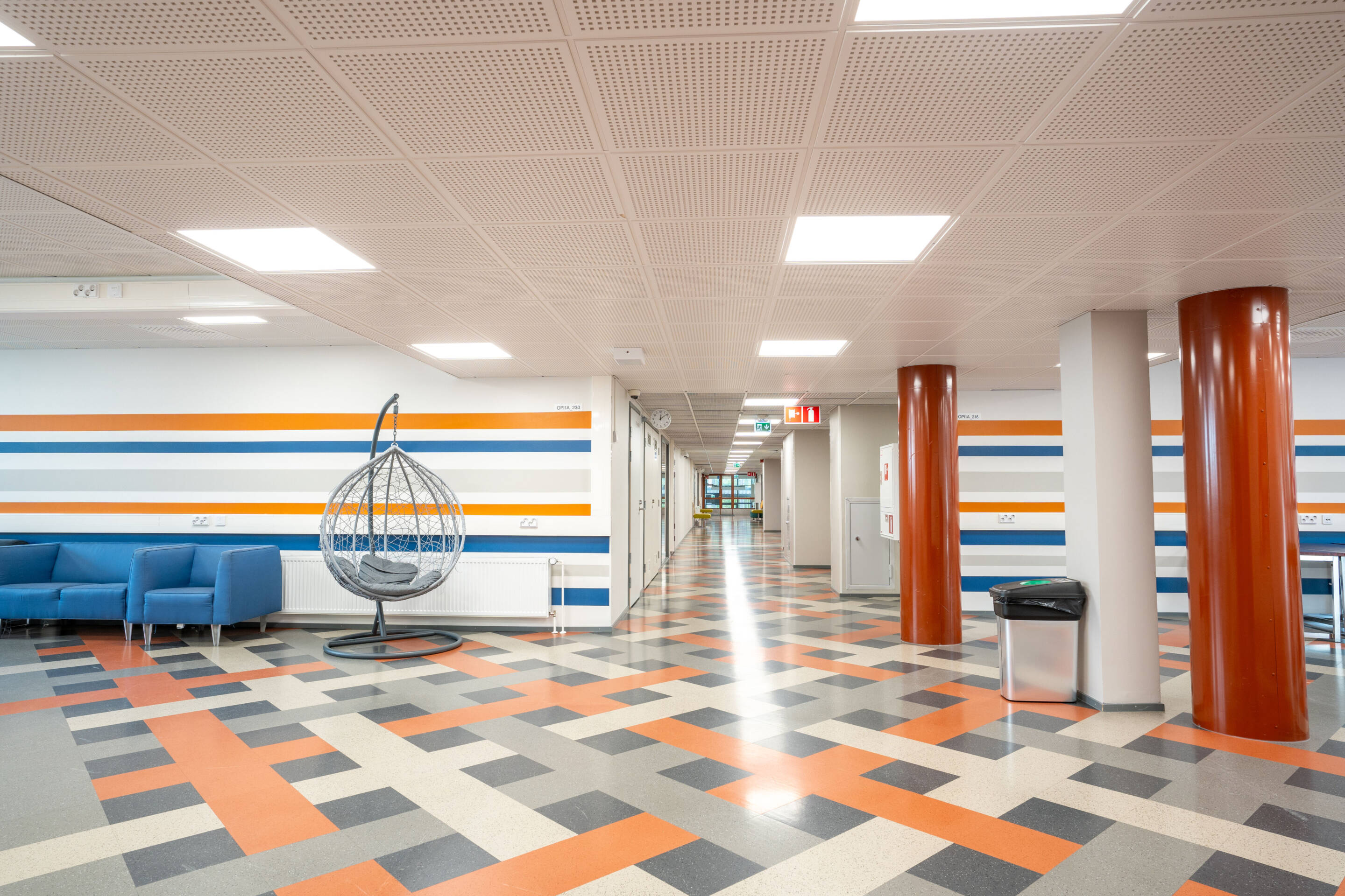 Moderni aula, jossa on geometrisesti kuvioitu lattiat ja joissa on sinisiä, oransseja ja valkoisia seinäraitoja. Vasemmalla on sininen sohva, jonka vierellä on harmaa riipputuoli ja kaksi pylvästä.