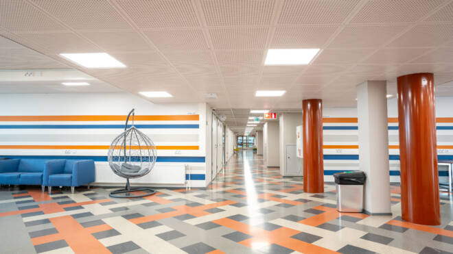 Moderni aula, jossa on geometrisesti kuvioitu lattiat ja joissa on sinisiä, oransseja ja valkoisia seinäraitoja. Vasemmalla on sininen sohva, jonka vierellä on harmaa riipputuoli ja kaksi pylvästä.