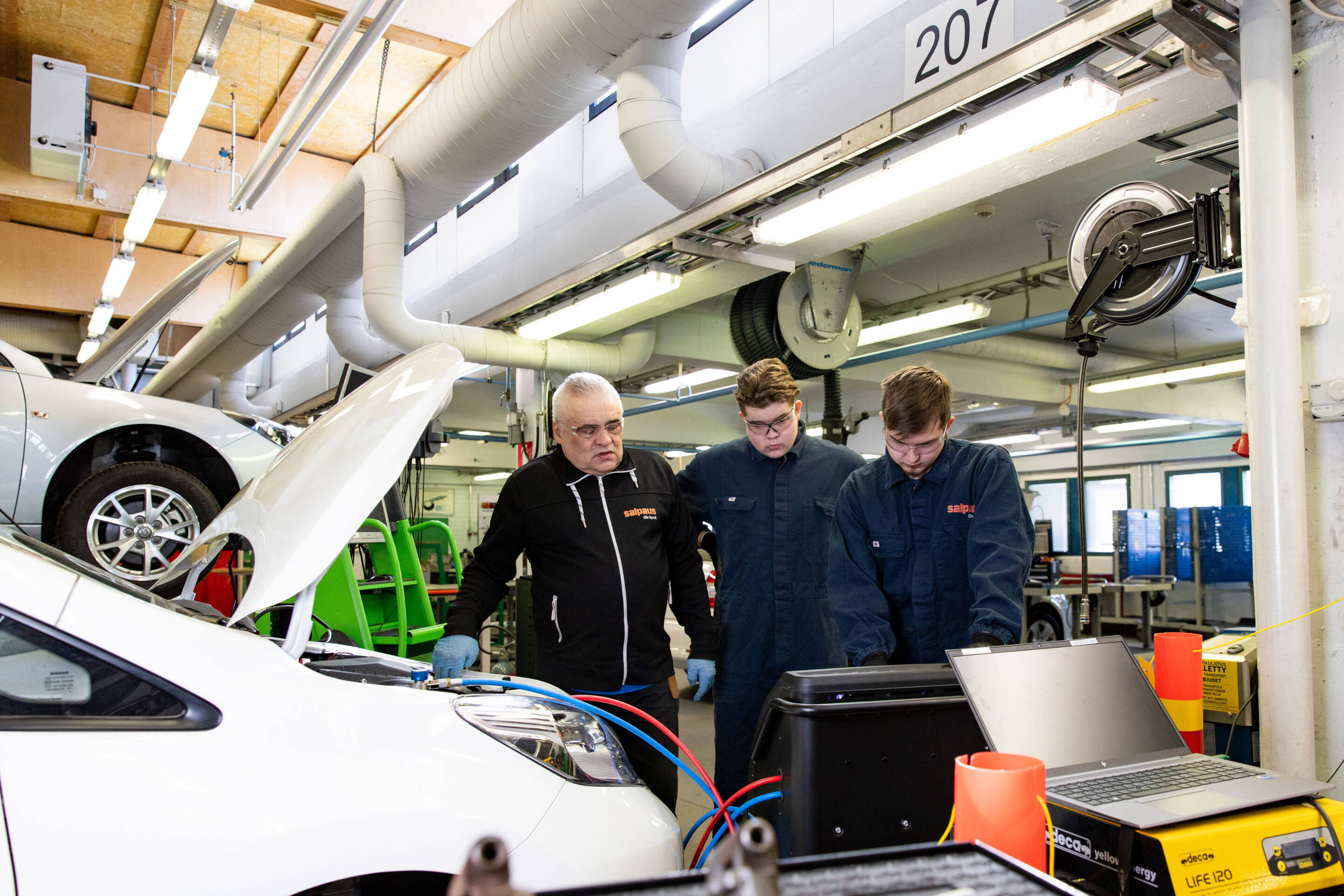 Kolmen hengen ryhmä seisoo valkoisen auton ympärillä autokorjaamossa. Kahdella on yllään sininen haalari ja yksi musta takki. Ne keskittyvät kaapeleihin ja autoon kytkettyyn laitteeseen, jossa tietokoneet ja laitteet ovat lähellä.