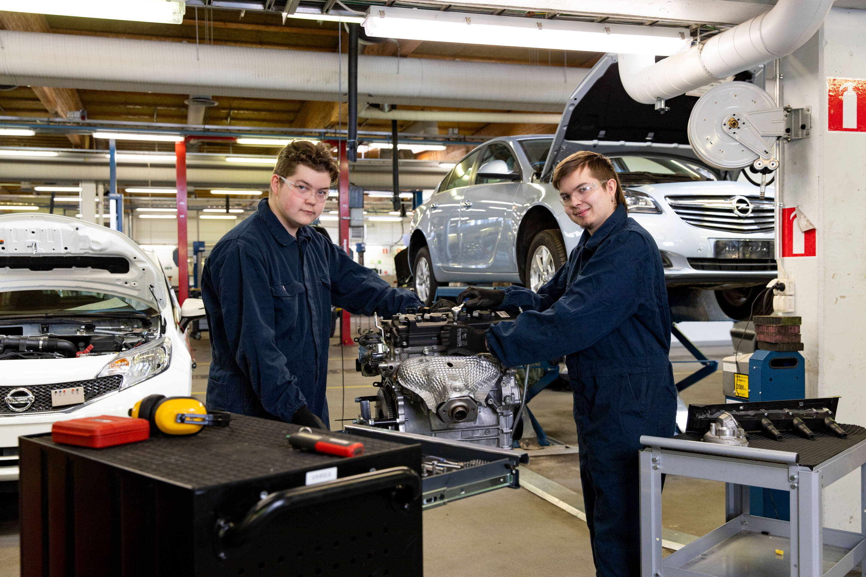 Kaksi mekaanikkoa sinisissä haalareissa työskentelee moottorin parissa autokorjaamossa. Yksi auto on hissillä ja toisen konepelti auki. .