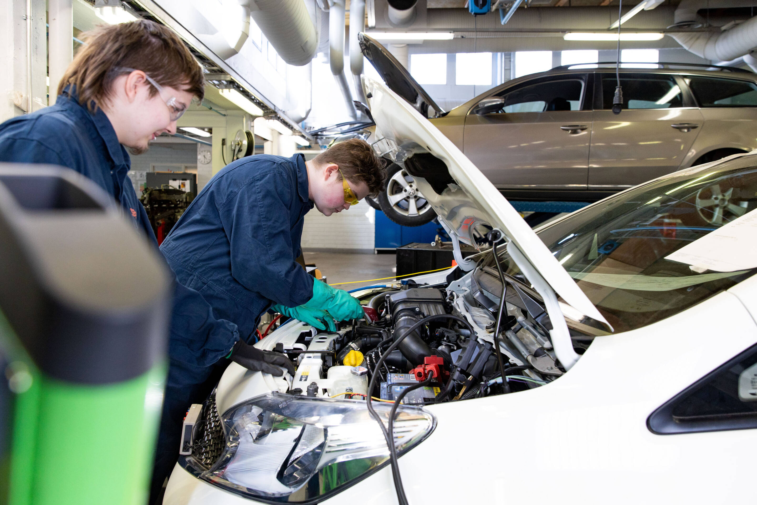 Kaksi mekaanikkoa sinisissä haalareissa työskentelee valkoisen auton moottorin parissa hyvin valaistussa autotallissa. Yksi kumartuu avoimen konepellin yli. Taustalla näkyy hopeanvärinen auto. Työkalut ja varusteet näkyvät.