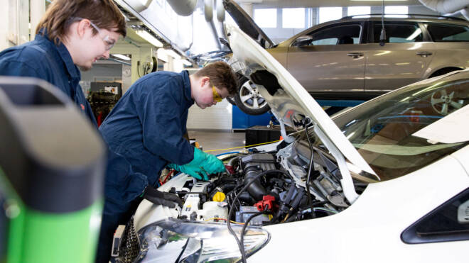 Kaksi mekaanikkoa sinisissä haalareissa työskentelee valkoisen auton moottorin parissa hyvin valaistussa autotallissa. Yksi kumartuu avoimen konepellin yli. Taustalla näkyy hopeanvärinen auto. Työkalut ja varusteet näkyvät.