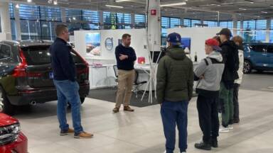 Ryhmä ihmisiä seisoo autonäyttelyssä ja kuuntelee jonkun puhuvan. Esillä on useita autoja, ja huoneessa on suuret ikkunat ja kirkas valaistus.