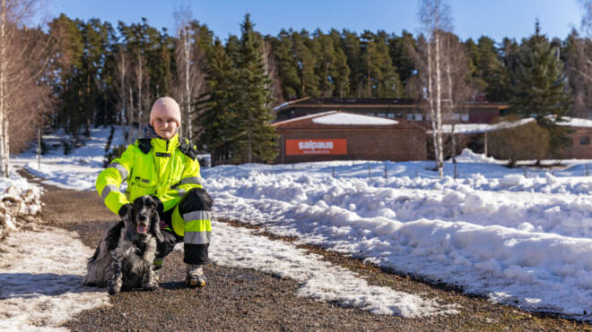 Kirkkaankeltaisessa takissa oleva henkilö polvistuu lumiselle polulle mustavalkoisen koiran viereen. Puut ja rakennus ovat taustalla, lunta hajallaan maassa. Taivas on kirkas ja sininen.