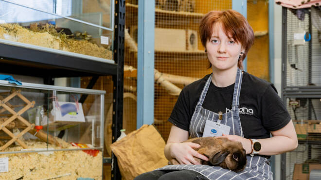 Lyhythiuksinen henkilö istuu häkeissä varustetussa huoneessa ruskea kani sylissään. Hänellä on yllään musta paita, raidallinen esiliina ja nimilappu. Taustalla näkyy erilaisia pieneläinten elinympäristöjä ja tarvikkeita.