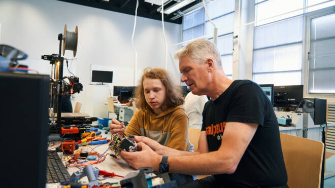 Vanhempi mies ja nuorempi työskentelevät yhdessä työpöydän ääressä, joka on täynnä elektroniikkaa ja johtoja tekniikan laboratoriossa. Mies selittää jotain pitäessään pientä laitetta. Nuorempi kuuntelee tarkkaavaisesti. Laitteet ja tietokoneet näkyvät taustalla.
