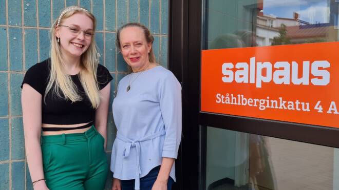Kaksi hymyilevää ihmistä lasioven vieressä, jossa oranssi kyltti, jossa lukee "salpaus, Ståhlberginkatu 4 A." Vasemmalla olevalla henkilöllä on yllään vihreät housut ja musta pusero, kun taas oikealla on vaaleansininen pusero.