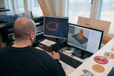 Henkilö työskentelee pöydän ääressä, jossa on kaksi tietokonenäyttöä. Näytöillä on 3D-mallinnusohjelmisto, joka näyttää yksityiskohtaisen mallin kohteesta. Näyttöjen vieressä on värikkäitä 3D-tulostettuja esineitä ja erilaisia pöytätyökaluja.
