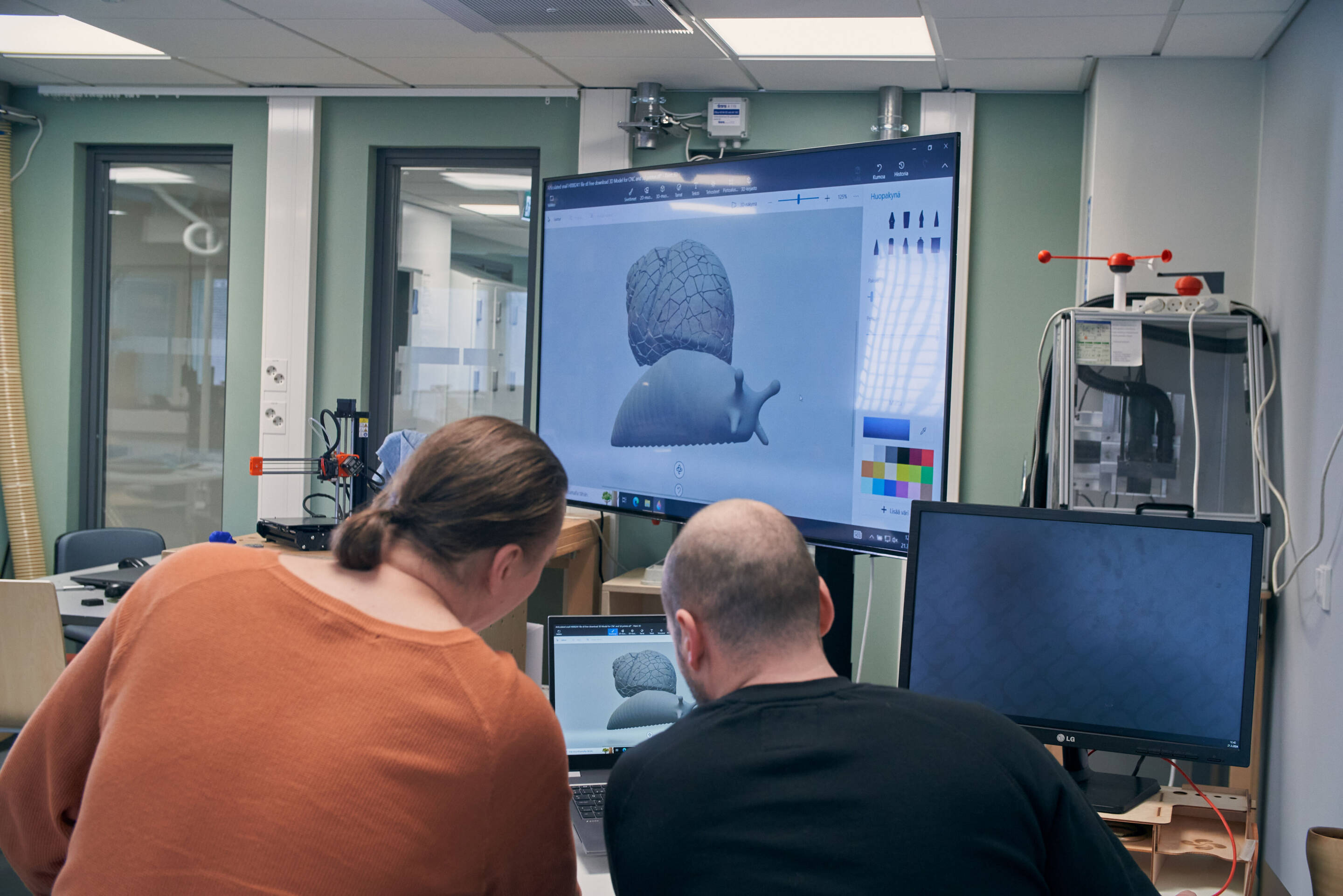 Kaksi ihmistä työskentelee digitaalisen 3D-mallin parissa suurella näytöllä modernissa toimistossa.
