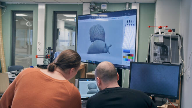 Kaksi ihmistä työskentelee digitaalisen 3D-mallin parissa suurella näytöllä modernissa toimistossa.