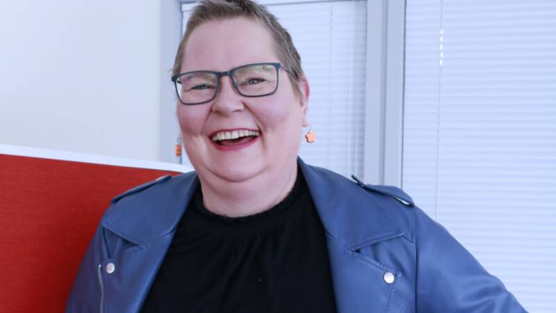 Lyhythiuksinen ja silmälasillinen henkilö seisoo hymyillen kädet lanteilla. Heillä on yllään sininen takki mustan mekon päällä sisätiloissa, joissa on valkoiset kaihtimet ja punainen väliseinä takana.