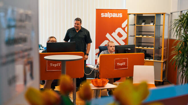 Toimisto, jossa kolme henkilöä työskentelee tietokoneiden ääressä. Mies seisoo ja kaksi muuta istuvat työpöydän ääressä, jonka oranssit taustat on merkitty TopIT-kylteillä. Huoneessa on hylly, kasvi ja roll-up, jossa on Salpaus. Tulppaanit ovat etualalla.