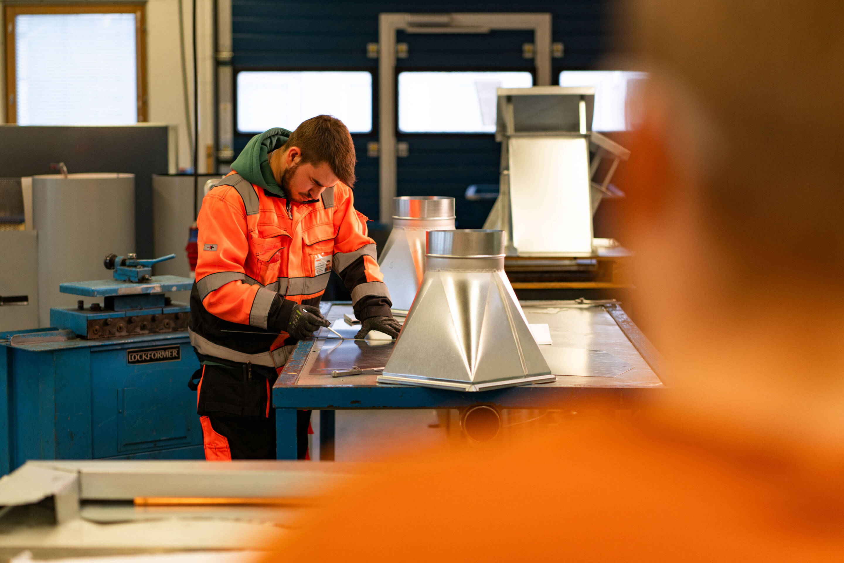 Oranssissa turvapuvussa oleva henkilö työskentelee metalliosien parissa. Koneet näkyvät taustalla. Etualalla näkyy hahmo samanlaisessa puvussa.