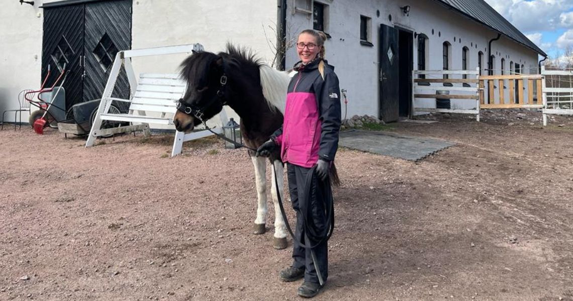 Nainen vaaleanpunaisessa ja harmaassa takissa seisoo pienen mustavalkoisen ponin vieressä tallin ulkopuolella. Talli on valkoinen ja mustat ovet. Kottikärryt ja valkoinen penkki ovat lähellä sorapolkua.