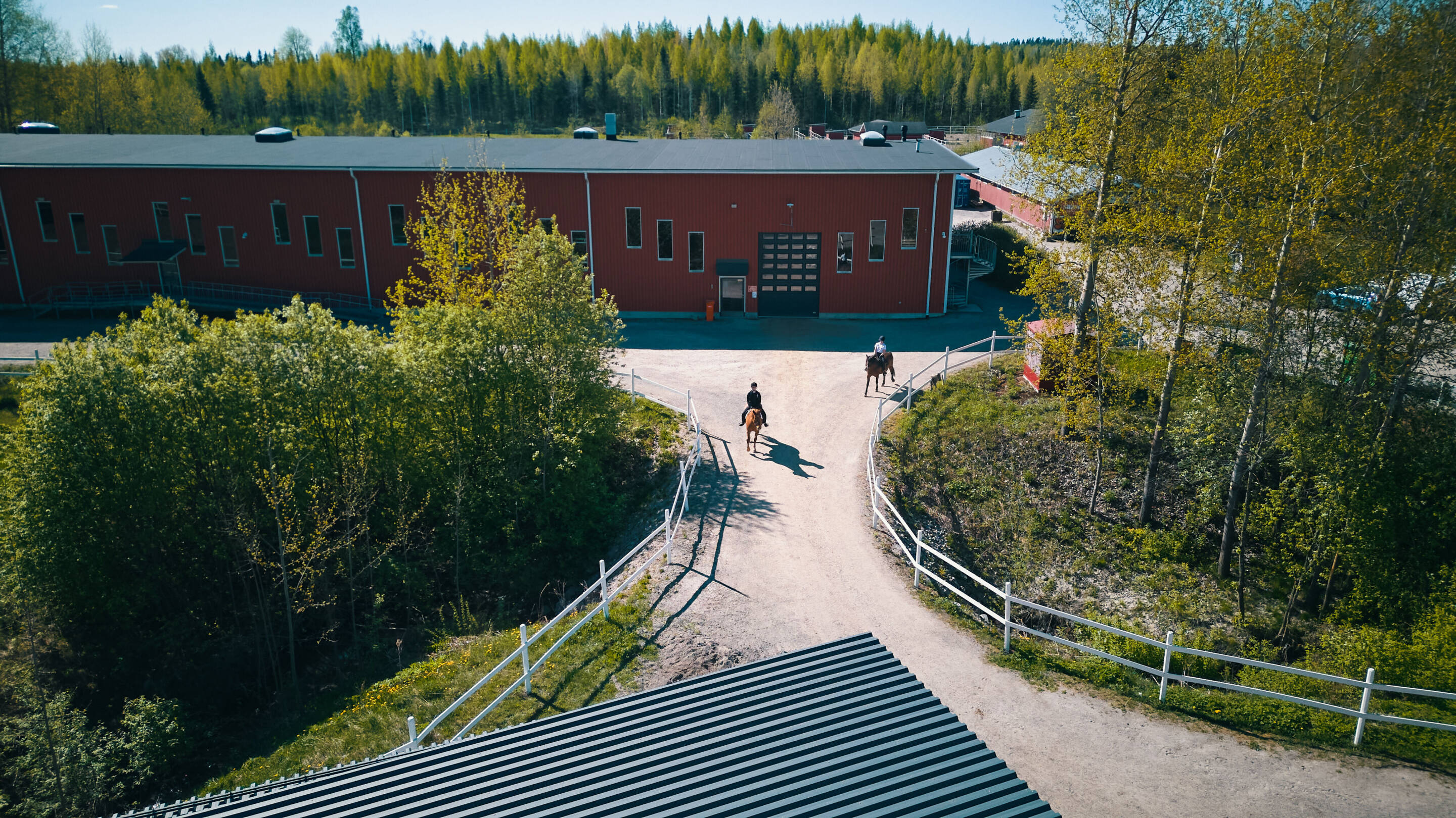 Ilmakuva navetta, jossa on punainen ulkopinta, puiden ympäröimänä. Kaksi ihmistä ratsastaa navettaan johtavalla hiekkapolulla. Aidat reunustavat polkua, ja maisema on vehreä ja metsä taustalla.