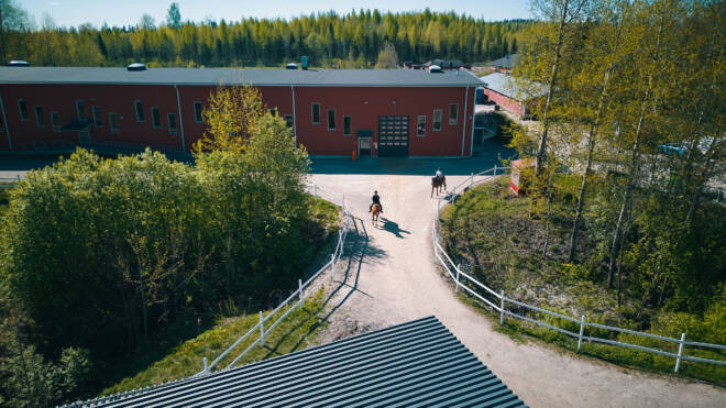 Ilmakuva navetta, jossa on punainen ulkopinta, puiden ympäröimänä. Kaksi ihmistä ratsastaa navettaan johtavalla hiekkapolulla. Aidat reunustavat polkua, ja maisema on vehreä ja metsä taustalla.