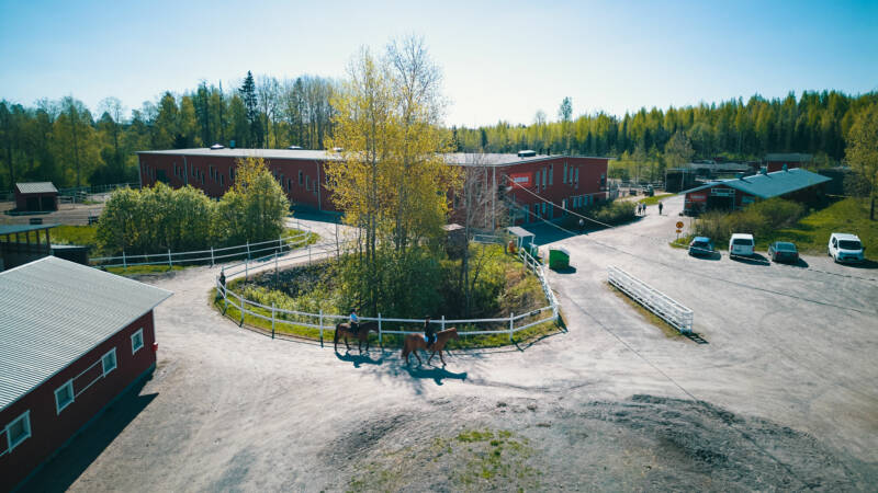 Ilmakuva hevosalan kampuksesta Lahden Jokimaalla, jossa on punaisia rakennuksia, puiden ympäröimänä aurinkoisena päivänä. Kaksi ihmistä ratsastaa hevosilla valkoisen aidan ympäröimää hiekkapolkua pitkin. Useita pysäköityjä autoja näkyy tien varrella lähellä rakennuksia.