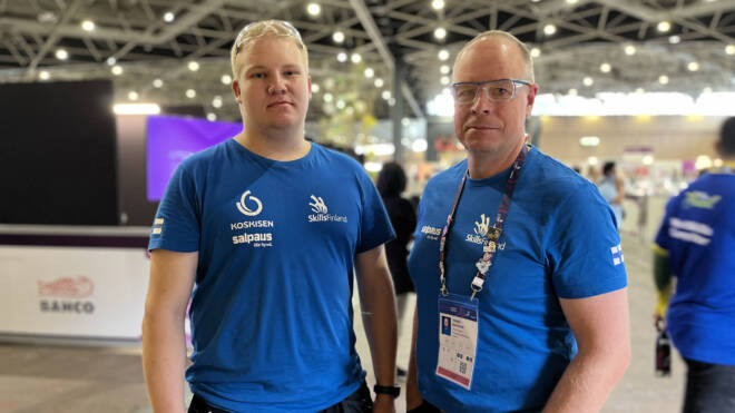 Kaksi miestä seisoo yhdessä sisätiloissa sinisissä Skills Finland -paidoissa. Vasemmalla miehellä on lyhyet vaaleat hiukset ja oikealla silmälasit ja kaulanauha, jossa on henkilökortti. Taustalla näkyy hyvin valaistu tapahtumatila.