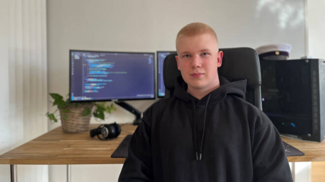 Nuori mies mustassa hupparissa istuu puisen pöydän edessä, jossa on kaksi näyttöä, joissa näkyy koodia. Ohjelmistokehittäjän takana työpöydällä on myös tietokone, kuulokkeet ja ylioppilaslakki.