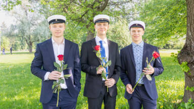 Kolme nuorta miestä puvuissa ja kaksoistutkintolakissa seisoo ulkona vehreässä puistossa. Jokaisella on punainen ruusu. He hymyilevät ja seisovat puun alla, jossa on vehreät lehdet.