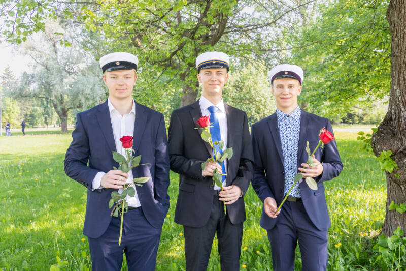 Kolme nuorta miestä puvuissa ja kaksoistutkintolakissa seisoo ulkona vehreässä puistossa. Jokaisella on punainen ruusu. He hymyilevät ja seisovat puun alla, jossa on vehreät lehdet.