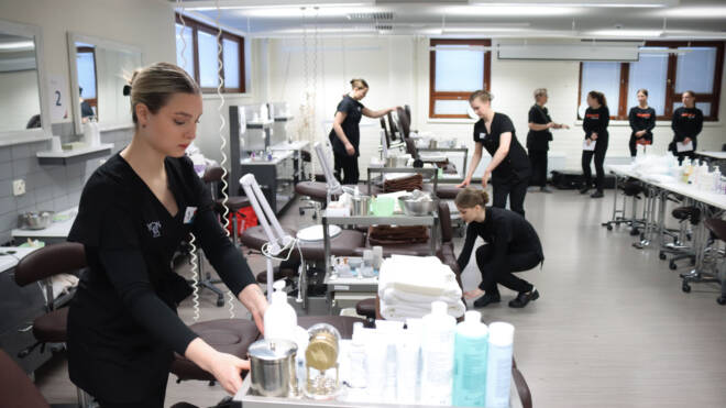 Ryhmä mustiin työvaatteisiin pukeutuneita ihmisiä valmistelee salonkia ja järjestää tarvikkeita. Huoneessa on useita hoitopisteitä, joissa on tuolit, peilit ja erilaiset ihonhoitotuotteet siististi järjestettyinä.