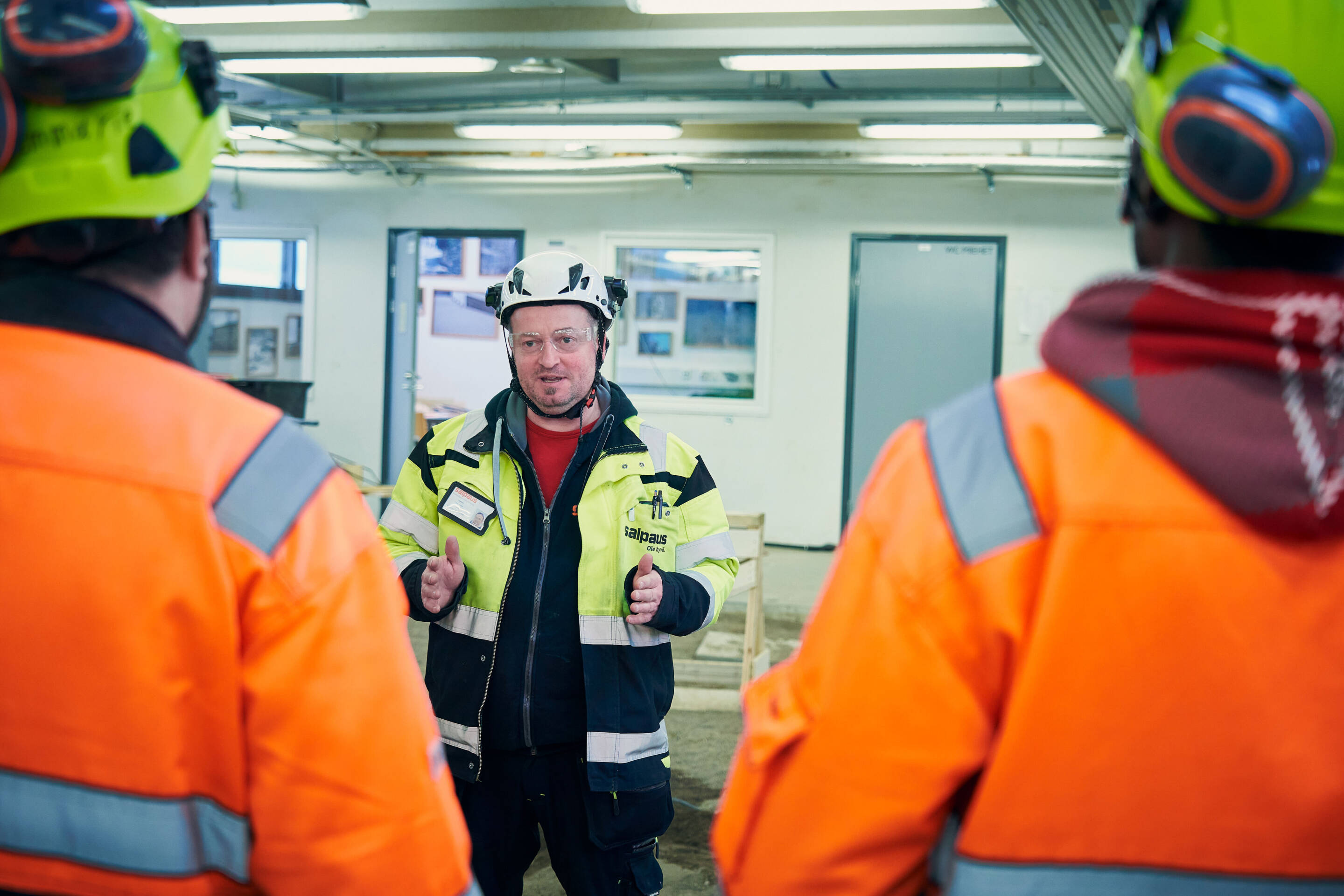 Turvavarusteissa oleva henkilö puhuu sisätiloissa kahdelle muulle kuvassa näkyvälle henkilölle, joilla on oranssit takit ja kypärät. Tapahtumapaikka näyttää olevan rakennus- tai teollisuusalue, jonka taustalla näkyvät loistelamput ja laitteet.