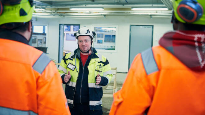 Turvavarusteissa oleva henkilö puhuu sisätiloissa kahdelle muulle kuvassa näkyvälle henkilölle, joilla on oranssit takit ja kypärät. Tapahtumapaikka näyttää olevan rakennus- tai teollisuusalue, jonka taustalla näkyvät loistelamput ja laitteet.