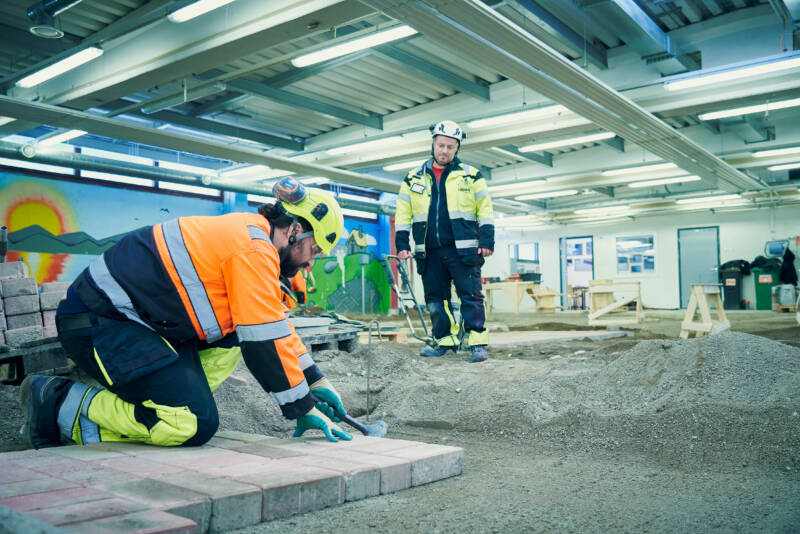 Kaksi rakennustyöntekijää turvavarusteissa työskentelee sisätiloissa. Toinen polvistuu maahan laskeen tiiliä, kun taas toinen seisoo tarkkailemassa. Tilavassa huoneessa on kirkkaat valot, hiekkainen maaperä ja värikkäitä seinämaalauksia seinillä.