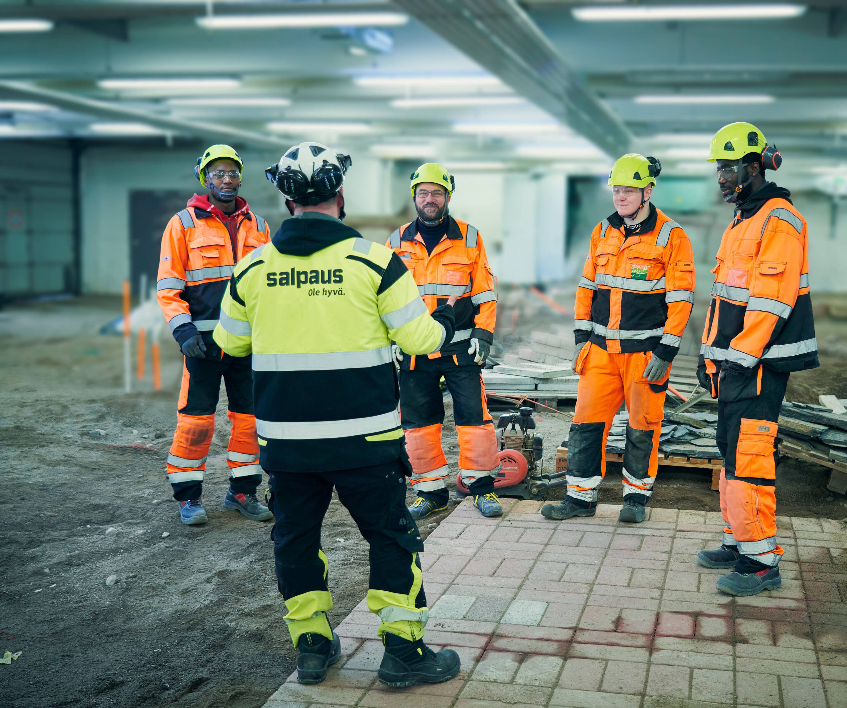Viisi rakennustyöntekijää, jotka pukeutuvat oranssiin ja mustiin näkyvyysvarusteisiin ja kypäriin, seisovat ympyrässä rakennustyömaalla.