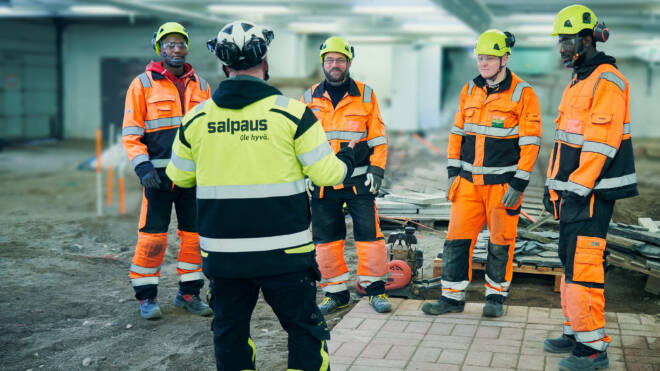 Viisi rakennustyöntekijää, jotka pukeutuvat oranssiin ja mustiin näkyvyysvarusteisiin ja kypäriin, seisovat ympyrässä rakennustyömaalla.