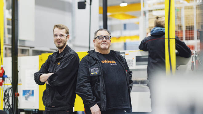 Kaksi mustaa univormua pukeutunutta miestä, mukaan lukien logolliset takit, seisovat selkä vastakkain hymyillen työpajassa. Koneet ja kirkkaan keltaiset aksentit näkyvät taustalla, mikä viittaa teolliseen ympäristöön.